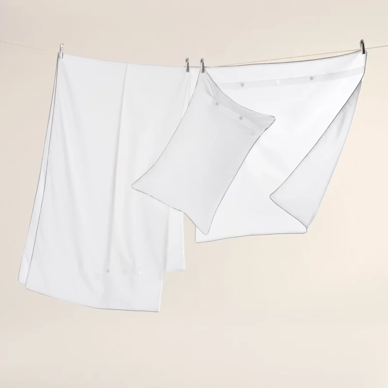 Bettwaesche weiss hängt auf einer Wäscheleine. Es ist ein Decken- und Kissenbezug sowie ein bezogenes Kissen zu sehen. Sie werden mit Metall Wäscheklammern festgehalten.