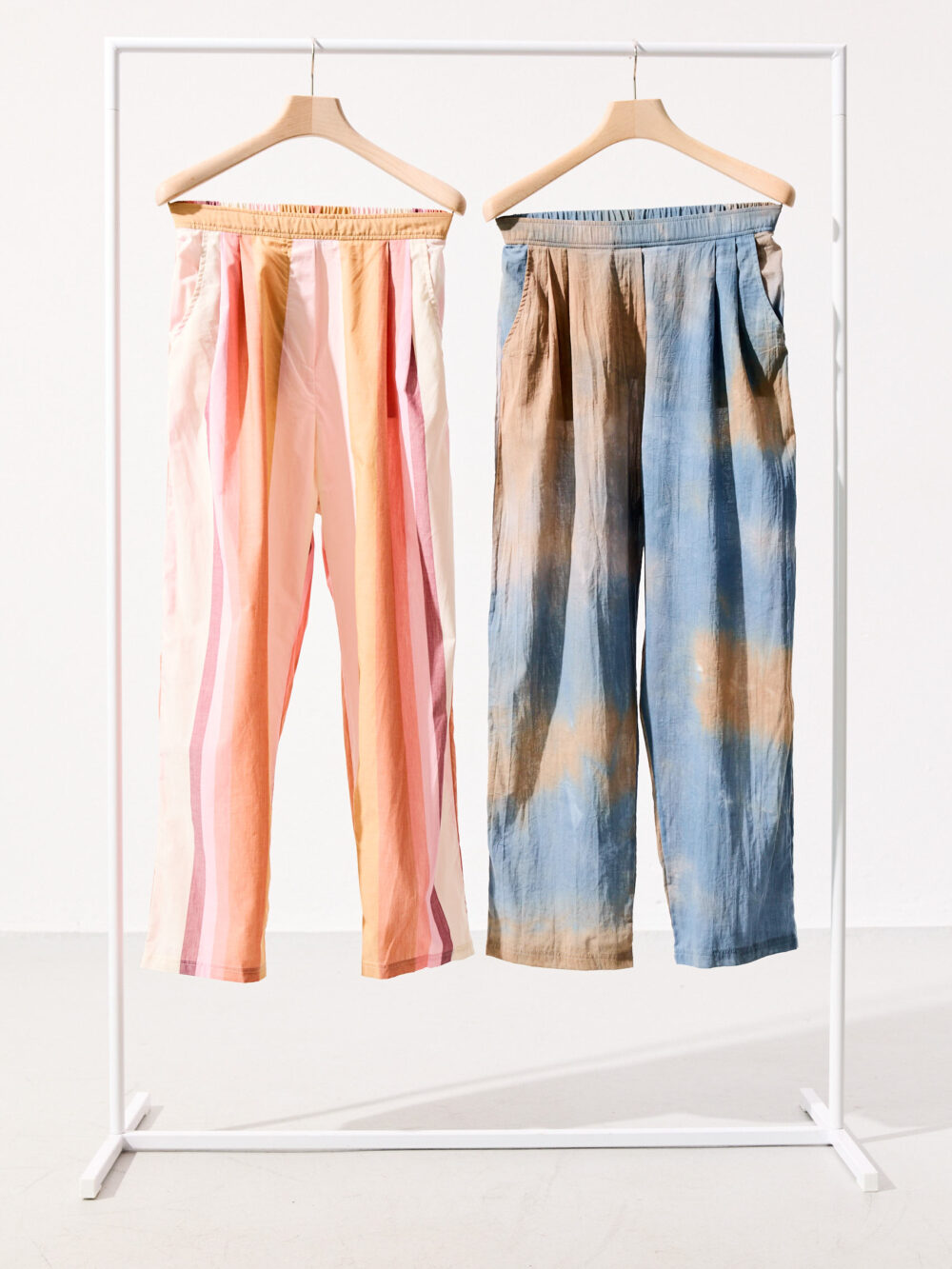 Stoffhose blau gebatikt hängt auf einem hellen Holzkleiderbügel an einer weißen Kleiderstange. Daneben die gleiche Hose in hellrosa gebatikt.