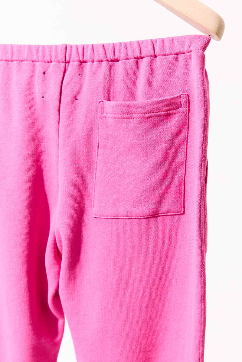 Detailaufnahme einer pinkfarbenen Sweathose mit elastischem Bund und aufgesetzter Gesäßtasche. Der weiche Baumwollstoff wirkt glatt gestrickt und elastisch, die Farbe leuchtet kräftig im Licht. Der obere Hosenbereich hängt locker auf einem Holzkleiderbügel vor hellem Hintergrund.
