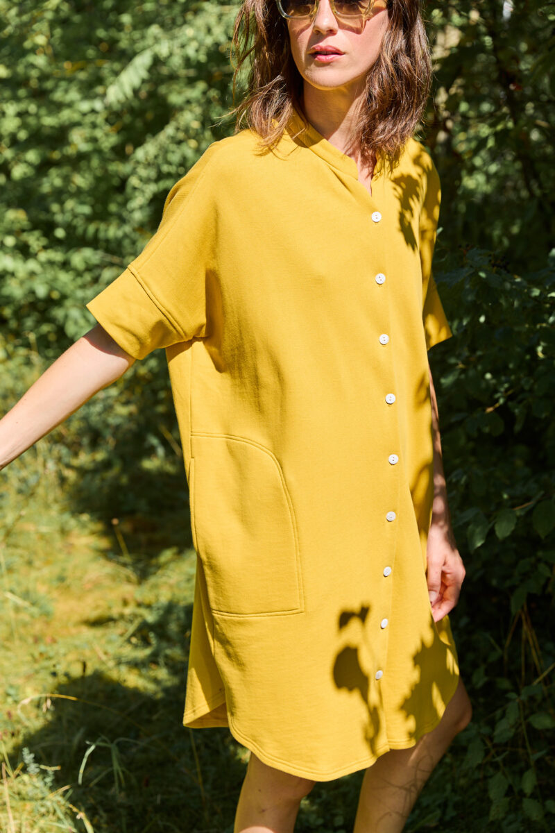 Lässiges Sweat Kleid in sun wird von einem stehendem Model, im Halbschatten getragen. Die Frau trägt eine Sonnenbrille und hat braune kurze Haare.